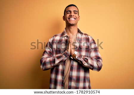 Young handsome african american man wearing casual shirt standing over yellow background praying with hands together asking for forgiveness smiling confident.
