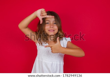 Little caucasian girl with blue eyes wearing white dress standing over isolated red background  making finger frame with hands. Creativity and photography concept.