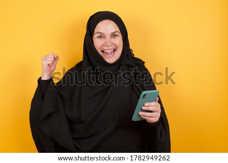 Beautiful middle aged muslim woman wearing black hijab over yellow background holding in hands cell celebrating 