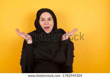 Surprised terrified Beautiful middle aged muslim woman wearing black hijab over yellow background Gestures with uncertainty, stares at camera, puzzled as doesn't know answer on tricky question, 