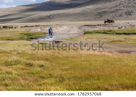 A motorcyclist tries to drive along a road filled with water during a flood after rain. Horses on the back plane.