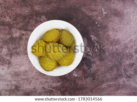 Top view of a small white bowl filled with hamburger dill pickle slices offset on a maroon tabletop illuminated with natural lighting.