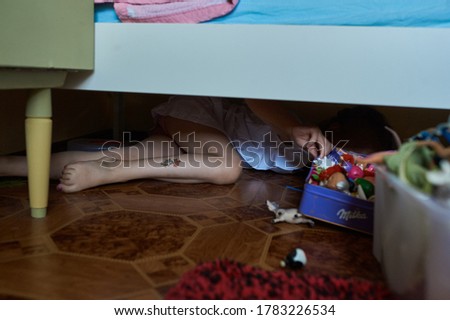A girl hiding under a bed with toys. Girl in white dress lies on the floor under the crib . High quality photo