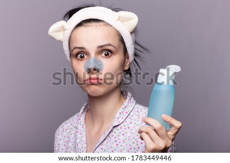 funny woman in a long-eared hair band holding a cosmetic bottle with a face tonic, a plaster from black dots on the nose, treatment of problem skin, gray studio background