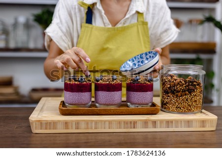 Woman making delicious desert chia puddings with strawberry and blueberry, almond milk with dragon fruit pink powder, and granola in kitchen at home.  
