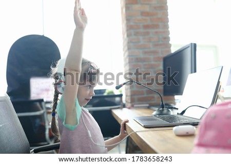 Little girl sit on chair and learn lesson online. Children master classe in video communication remotely. Streaming and games online. Child look in headphones at laptop and hold hand up. Training for