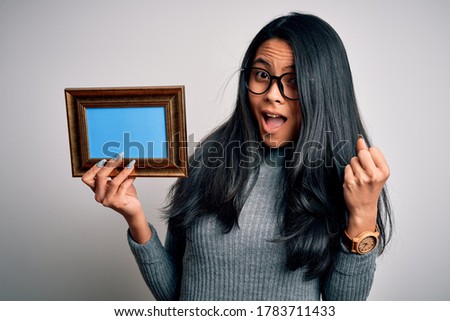 Young beautiful chinese woman holding vintage frame over isolated white background screaming proud and celebrating victory and success very excited, cheering emotion