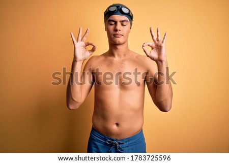 Young handsome man shirtless wearing swimsuit and swim cap over isolated yellow background relaxed and smiling with eyes closed doing meditation gesture with fingers. Yoga concept.