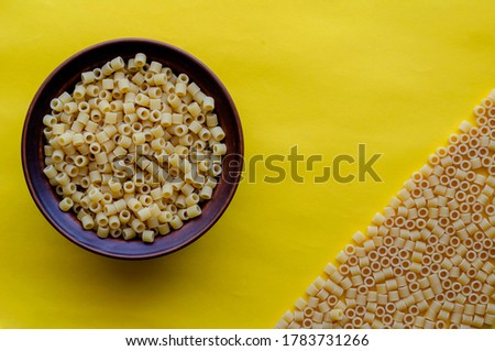 Clay bowl with ditalini pasta on a yellow background. The right corner is occupied by dry ditalini. Variety of Italian pasta. View from above. Copy space.