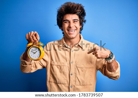 Young handsome man holding alarm clock standing over isolated blue background with surprise face pointing finger to himself