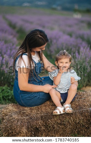 Mom and little daughter sits on hay by the farm. Background of summer lavender field. Family denim style