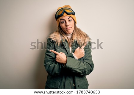 Young brunette skier woman wearing snow clothes and ski goggles over white background Pointing to both sides with fingers, different direction disagree
