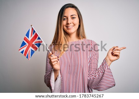 Young beautiful blonde woman with blue eyes holding united kingdom flag very happy pointing with hand and finger to the side