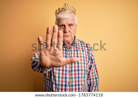 Senior handsome hoary man wearing golden crown of king over isolated yellow background doing stop sing with palm of the hand. Warning expression with negative and serious gesture on the face.