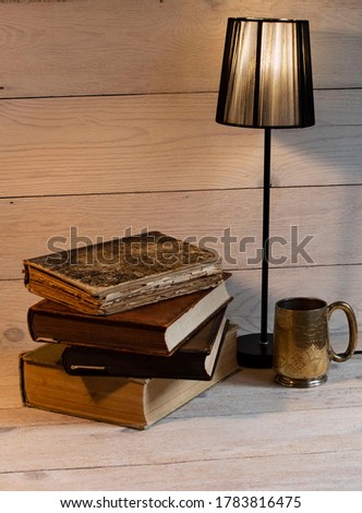 Still life from a stack of old books, a night lamp and a mug of tea, on a light wooden table.