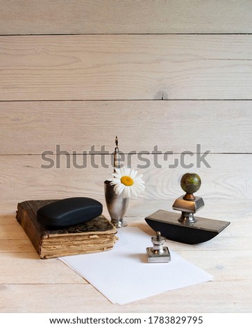 Old book, silver writing set of ink pens, paperweight, stamp and glass on a light table with white sheet of paper and chamomile Royalty-Free Stock Photo #1783829795