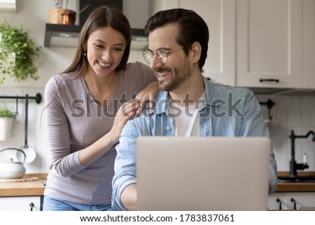Happy young Caucasian couple sit in kitchen look at laptop screen browsing internet surfing web together, smiling millennial husband and wife shopping online on computer at home, use gadget technology