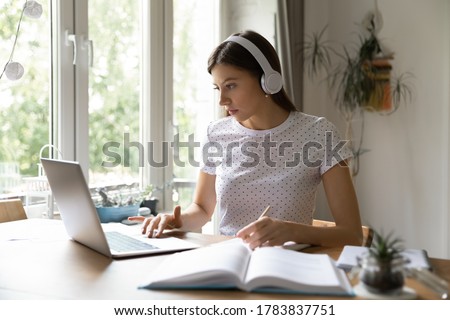 Young female student in earphones sit at desk at home look at laptop screen studying making notes, concentrated Caucasian girl in headphones watch webinar on computer, distant education concept Royalty-Free Stock Photo #1783837751