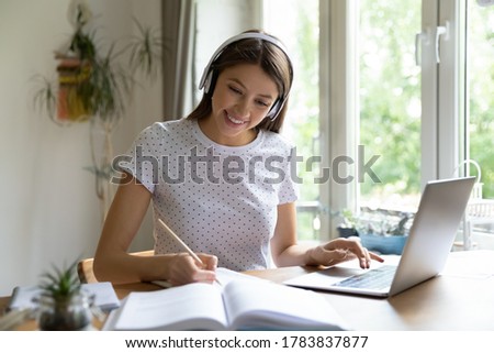 Smiling millennial Caucasian girl in headphones sit at desk study make notes using laptop, happy young female student in earphones make homework, take online course, distant education concept Royalty-Free Stock Photo #1783837877