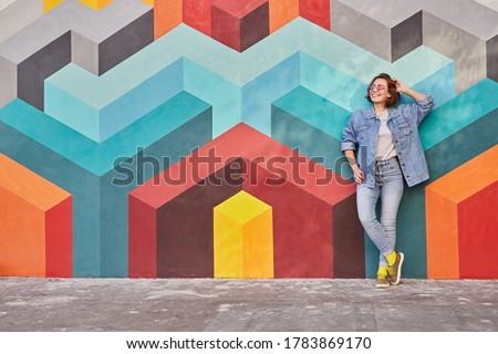Relaxed young female in denim outfit smiling and looking away while leaning on multicolored graffiti wall with geometric ornament on city street Royalty-Free Stock Photo #1783869170