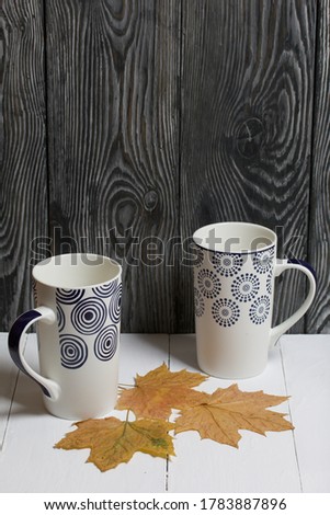Ceramic mug on a white surface. Between them are dried autumn maple leaves. Against a background of pine boards painted black.