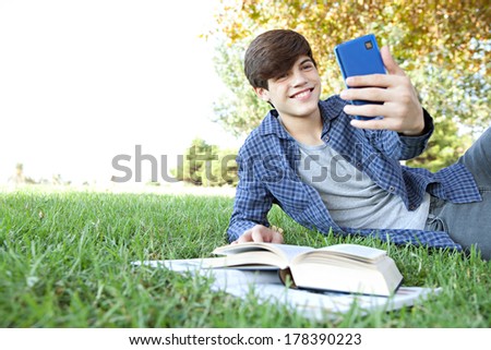 Portrait view of a teenager student boy laying down in a park with his college books, using a smartphone to take a selfie photo of himself for networking, posing and smiling. Technology lifestyle.