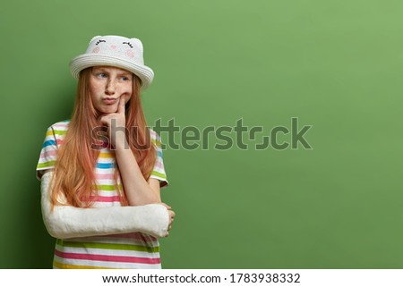Pensive freckled girl with ginger hair, keeps finger on cheek, has displeased expression, has broken arm, cannot play with children outdoor, isolated on green background, empty space for promo