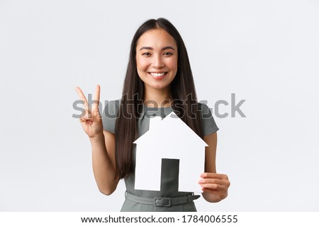 Insurance, loan, real estate and family concept. Smiling attractive asian woman selling or buying apartment, showing peace kawaii sign and paper house, saleswoman showing home, white background