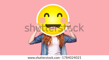 Redhead girl hiding her face behind happy emoji smile, posing over pink studio background, panorama with copy space Royalty-Free Stock Photo #1784024021