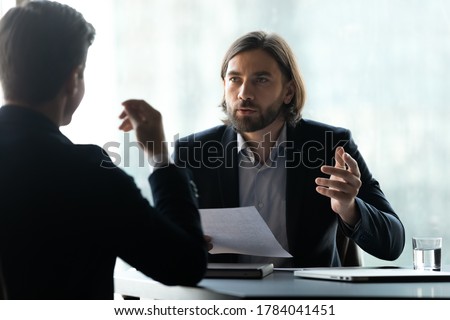 Serious young executive manager with long hairstyle in formal suit discussing business agreement details with focused male partner at negotiation meeting, hr holding job interview with candidate.