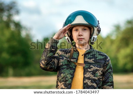 A boy in a pilot's helmet on a background of greenery salutes. Dream concept, choice of profession, military service. Copy space