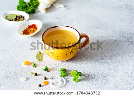 Homemade chicken (bone) broth/bouillon with vegetables, spices and herbs in a bowl (cup) on a light background. Natural collagen of animal origin. Liquid broth from meat, fish, vegetables.