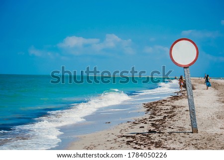 Seaside with danger sign, white middle and red edges, like warning about next steps. Dangerous zone for walking and swimming. Coastline with bright blue water and sand beach.