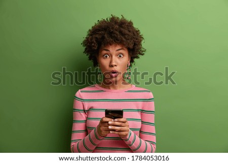 Cute astonished woman uses online application, uses cell phone for chatting in social networks, stunned to read amazing news, shocked about received message, stands indoor. Technology concept