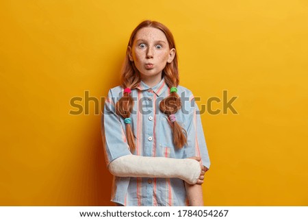 Surprised preteen girl pouts lips and looks with bugged eyes at camera, makes funny grimace and foolishes around, wears gypsum on broken arm after accident on road. Children, face expressions