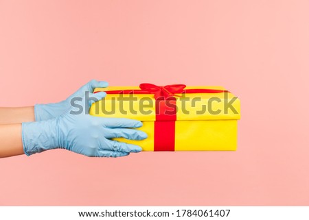 Profile side view closeup of human hand in blue surgical gloves holding yellow gift box. indoor, studio shot, isolated on pink background.