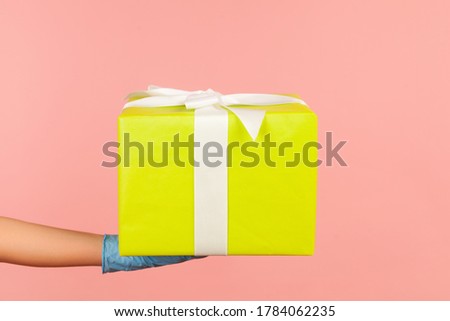 Profile side view closeup of human hand in blue surgical gloves holding gift box. sharing, giving or delivery concept. indoor, studio shot, isolated on pink background.
