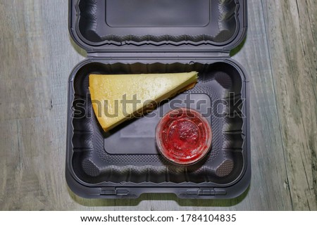 A take out container with a piece of cheesecake and disposable sauce cup. 