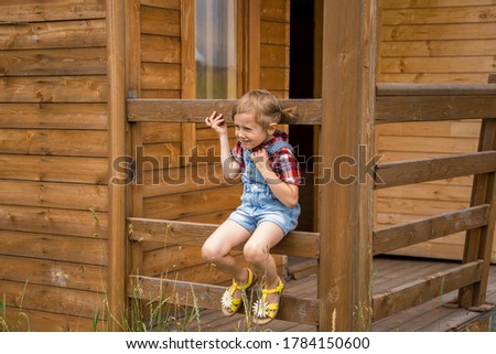 Adorable Children girl, kid cowgirl sitting on a fence of a wooden house. happy childhood in a village