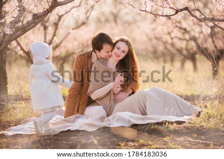 Happy family are relaxing on a picnic in the blooming peach gardens at sunset in the sun. Mother and father hug, kiss their little daughter, sit on a blanket, laugh. A weekend trip with children 