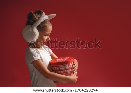 Merry Christmas! happy little girl in fur headphones with a magic gift on a dark red background. The child enjoys the holiday. christmas present. Advertising. Christmas eve and spirit