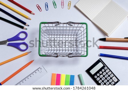 Top view photo of a small shopping basket in the center and colorful office supplies, isolated on a white background