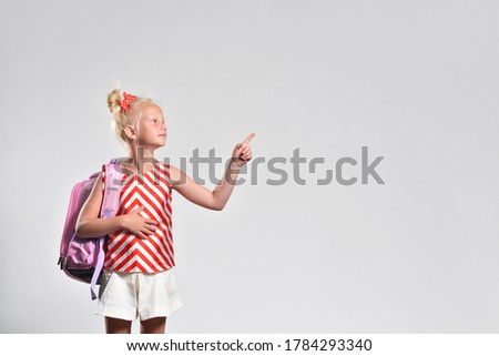 Schoolgirl with backpack points finger at text space on gray background, studio photography.