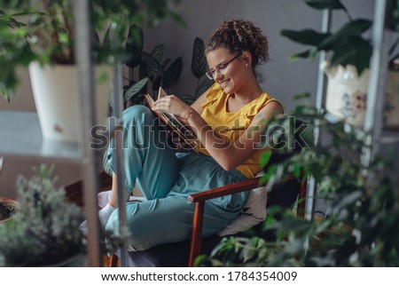 Woman reading a book in room of plants. Young florist in eyeglasses sitting on armchair and reading a book.