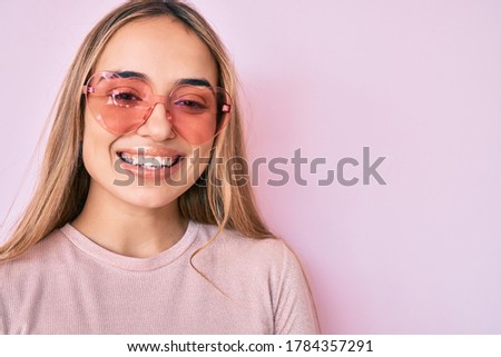 Young beautiful blonde woman wearing heart shaped sunglasses smiling with a happy and cool smile on face. showing teeth. 