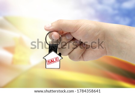 A man's hand holds a keychain in the form of a house with the inscription Sale on the background of the flag of Zimbabwe. Property and real estate sale concept.