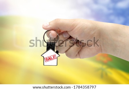 A man's hand holds a keychain in the form of a house with the inscription Sale on the background of the flag of Saskatchewan. Property and real estate sale concept.