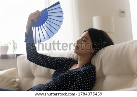 Unwell millennial Asian girl relax on sofa in living room wave with hand fan suffer from hot weather, have no air conditioner, overheated Vietnamese woman use waver struggle with hormonal imbalance Royalty-Free Stock Photo #1784375459