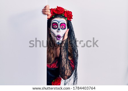 Young woman wearing day of the dead custome holding blank empty banner afraid and shocked, surprise and amazed expression with hands on face 
