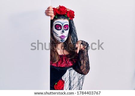 Young woman wearing day of the dead custome holding blank empty banner pointing down looking sad and upset, indicating direction with fingers, unhappy and depressed. 